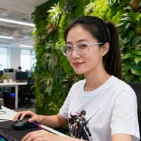 一位约28岁的年轻女性用户头像照片，扎着马尾辫，戴着银色细框眼镜，身穿白色T恤，背景为开放式办公室的绿植墙，她是一名电竞爱好者